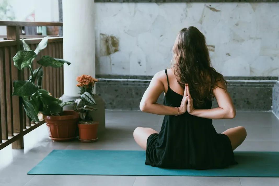 Woman seen from behind sitting on a teal mat with her hands in prayer position on a balcony with plants
