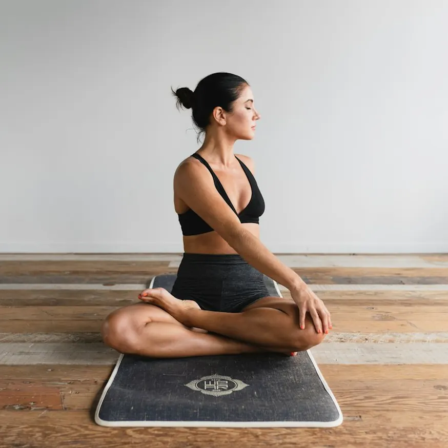 Woman in a black sports bra and shorts sitting cross-legged on a mat against a plain wall